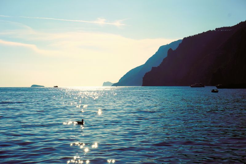Serene view of Amalfi Coast waters at sunset with distant boats and silhouetted cliffs.