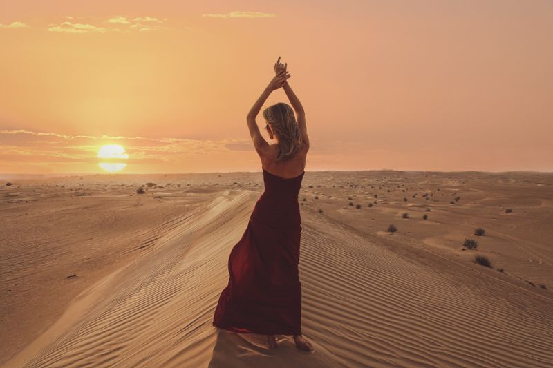 A woman in a red dress poses in the Dubai desert during a stunning sunset with sand dunes stretching into the horizon.