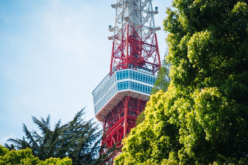 A striking view of Tokyo Tower rising above lush green trees under a bright blue sky.