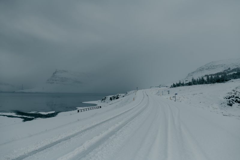 A serene winter landscape in Iceland featuring a snow-covered road alongside a tranquil fjord.