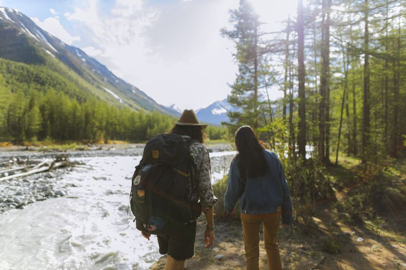 Couple hiking through a forest trail by a mountain river under bright sunlight, experiencing nature's beauty.
