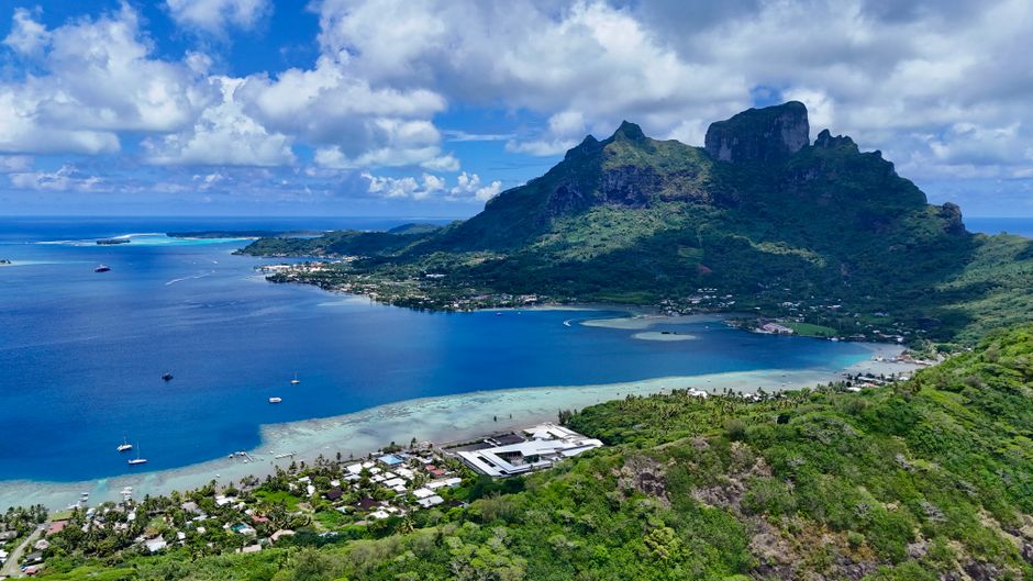Aerial view of Bora Bora's stunning lagoon and lush mountains under a bright blue sky.